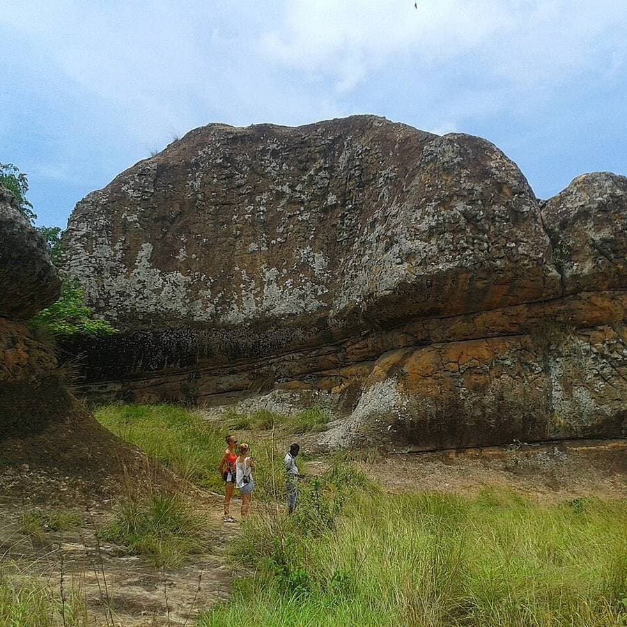 Énorme bloc de grès strié dominant une vallée herbeuse à Tanoboase, avec trois promeneurs minuscules à son pied