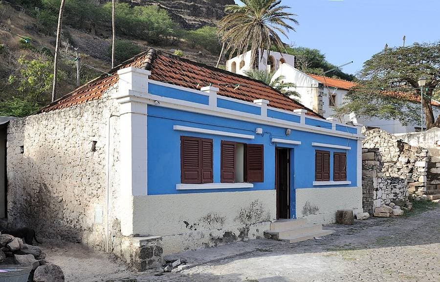 Maison basse aux murs bleus et volets bruns dans une rue pavée, une colline et des palmiers derrière