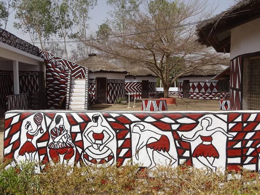 Muret peint de personnages et de chevrons rouges, noirs et blancs devant les cases rondes à toit de chaume de la SWOPA, à Sirigu