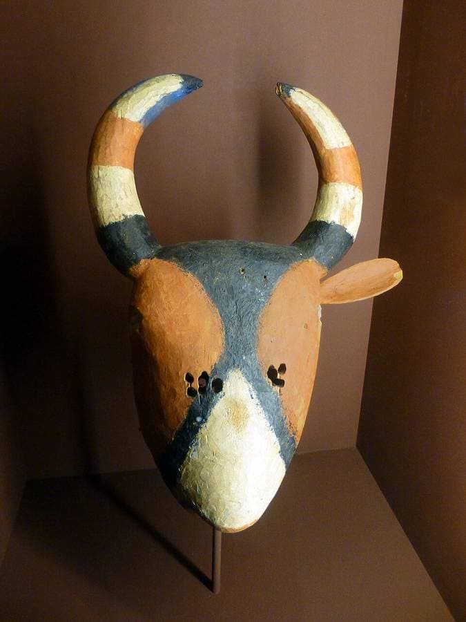 Masque de bovin en bois peint en ocre, bleu et blanc, cornes dressées, exposé sur un socle au Musée africain de Lyon