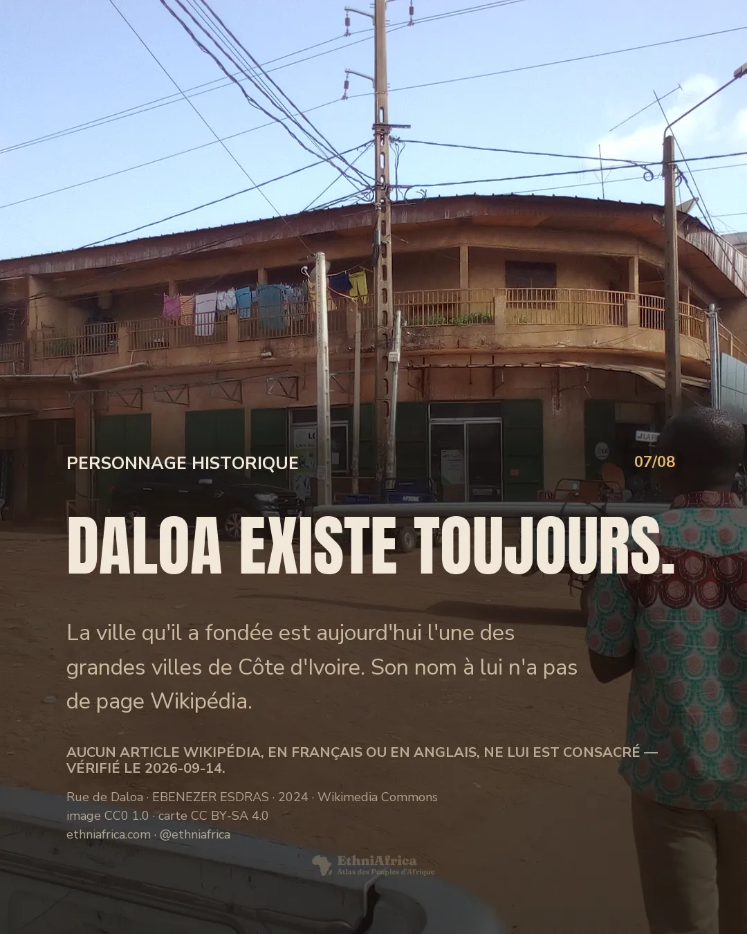 une rue en terre ocre d'un centre-ville, un immeuble à balcon incurvé avec du linge séchant, une voiture noire garée devant une boutique aux volets verts, un jeune homme de dos portant une barre de métal sur l'épaule au premier plan