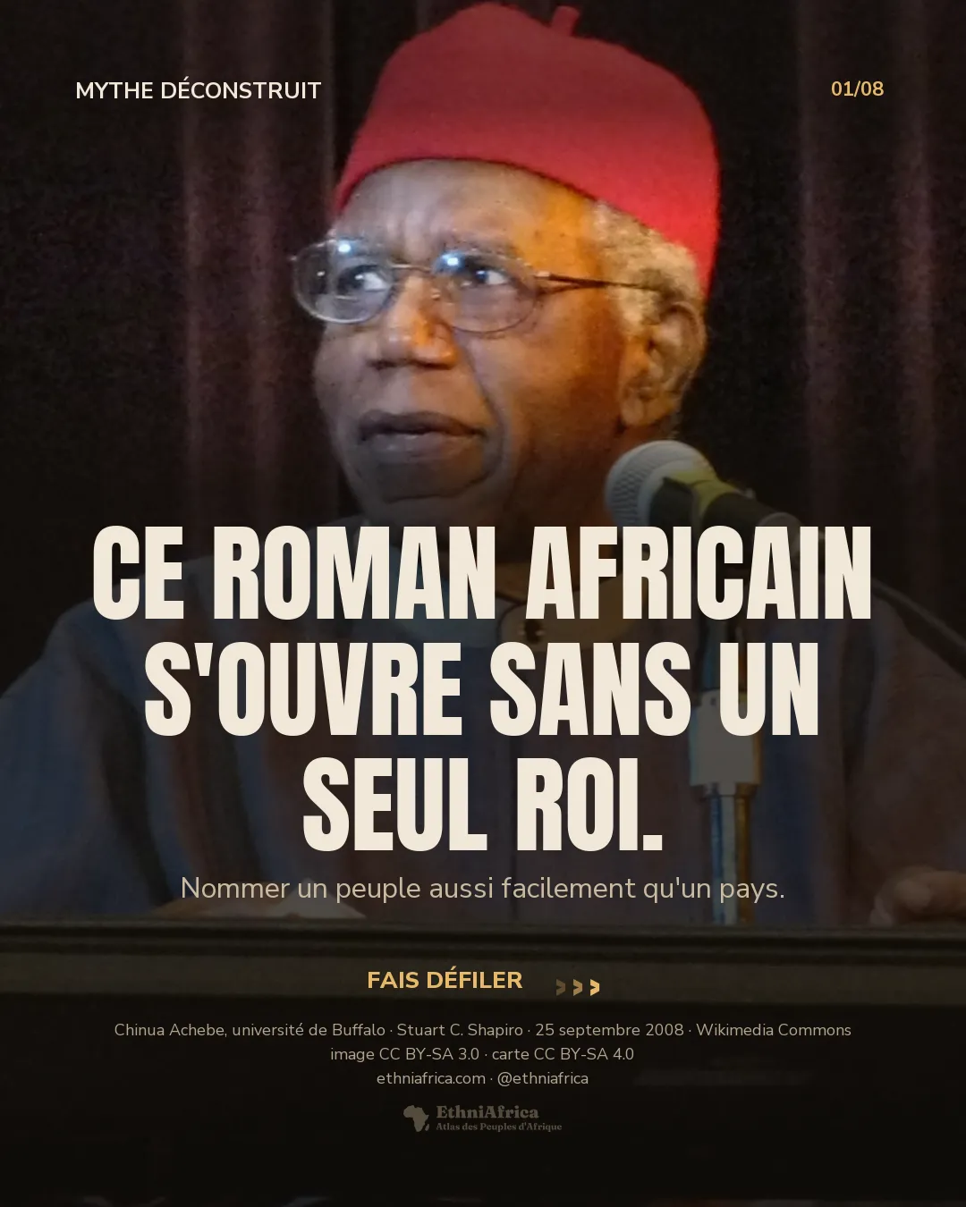 un homme âgé à lunettes, coiffé d'un bonnet rouge traditionnel igbo (okpu ozo), parle derrière un pupitre et un micro sur une scène, devant un rideau sombre