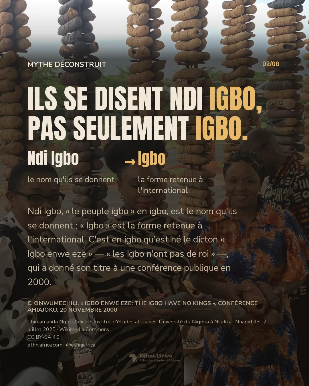 une femme au chignon tressé relevé, vêtue d'un imprimé zébré, sourit en tenant des ignames entre deux autres femmes, devant une installation faite de perles et de gourdes empilées sur des poteaux, en plein air