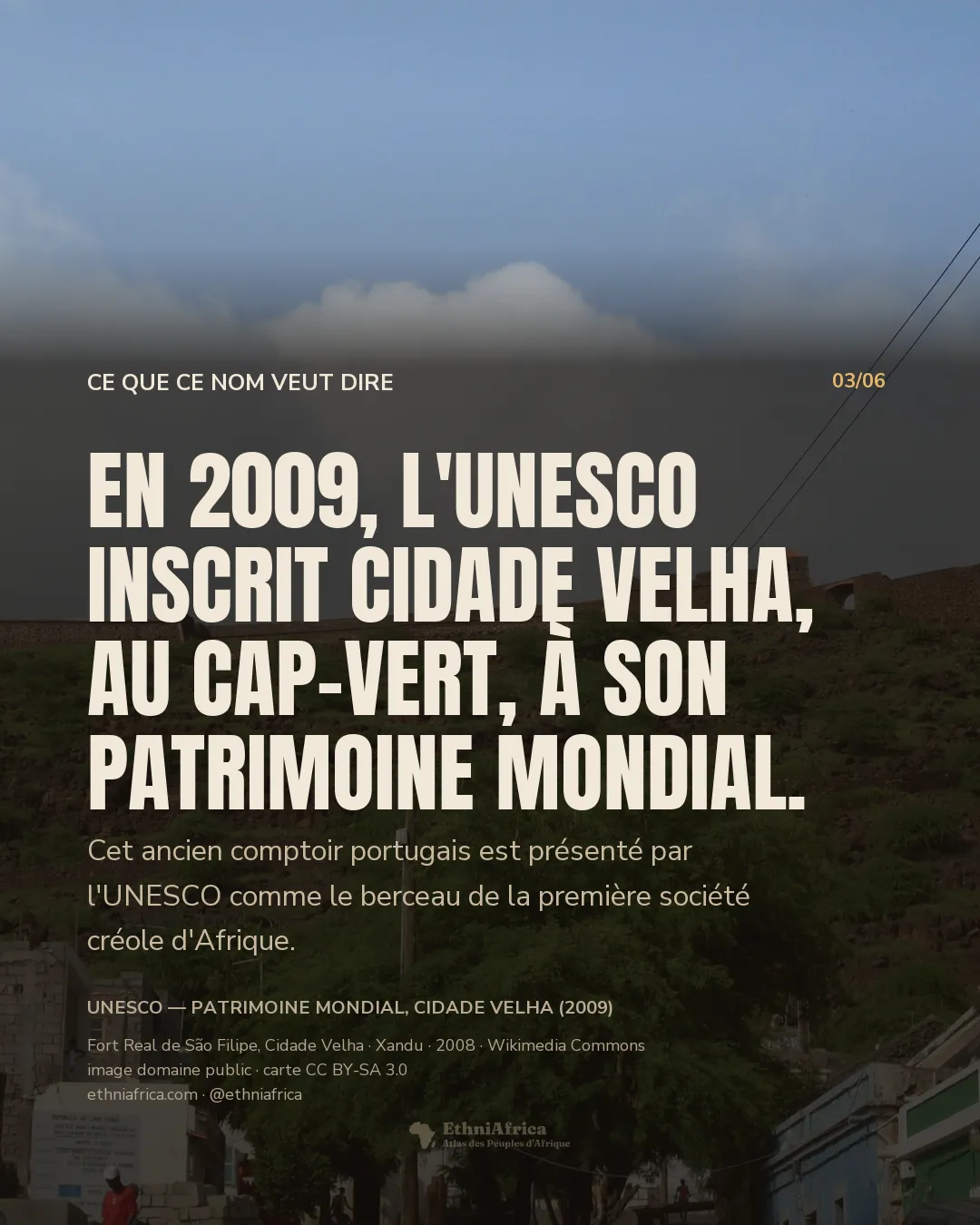 un mur de fortification en pierre couronnant une crête volcanique verte, vu depuis un village aux toits de tôle et de tuile en contrebas, sous un ciel bleu partiellement nuageux